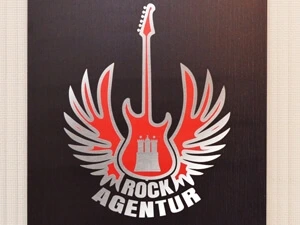 Schwarzes Schild der Rockagentur mit roten Logo und silbernen Outlines.