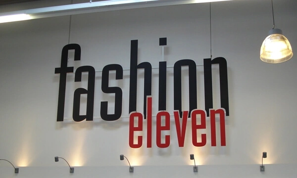 Logo eines Textilhandels aus Fräsbuchstaben in Rot und Schwarz an einer Wand, beleuchtet von Strahlern