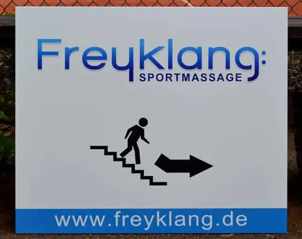 Montiertes Schild mit der Aufschrift "Freyklang Sportmassage" und einem Männchen auf einer Treppe mit Pfeil als Wegweiser.