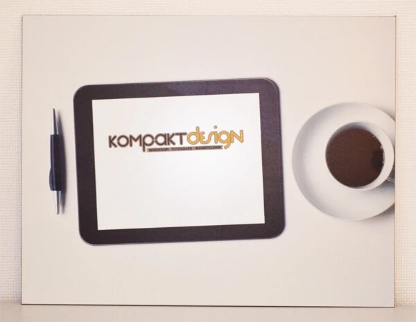 Ein Schild mit Foto als Direktdruck - Heller Hintergrund mit Kugelschreiber, Tablet auf dem das Kompaktdesign-Logo zu sehen ist und eine Tasse mit Kaffee.
